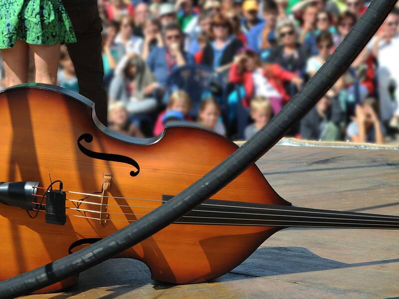 Ein Cello Liegt auf einer Bühne. Im Hintergrund sind Zuschauerinnen und Zuschauer erkennbar. 