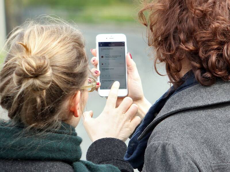 Zwei Frauen sind von hinten zu sehen und schauen gemeinsam auf ein Smartphone, auf dem die Nina-Warnapp gezeigt wird. 