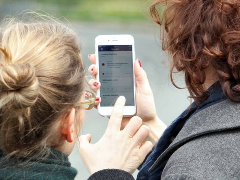 Zwei Frauen sind von hinten zu sehen und schauen gemeinsam auf ein Smartphone, auf dem die Nina-Warnapp gezeigt wird. 
