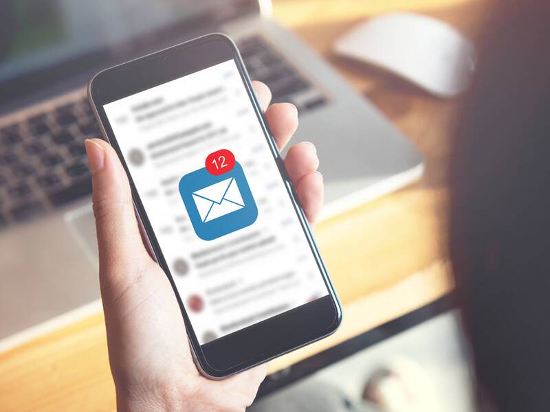 Eine weibliche Person, von hinten fotografiert, hält ein Handy in der Hand, auf dem ein E-Mail-Symbol aufleuchtet. Im Hintergrund ist zudem ein offenes Notebook zu sehen. 