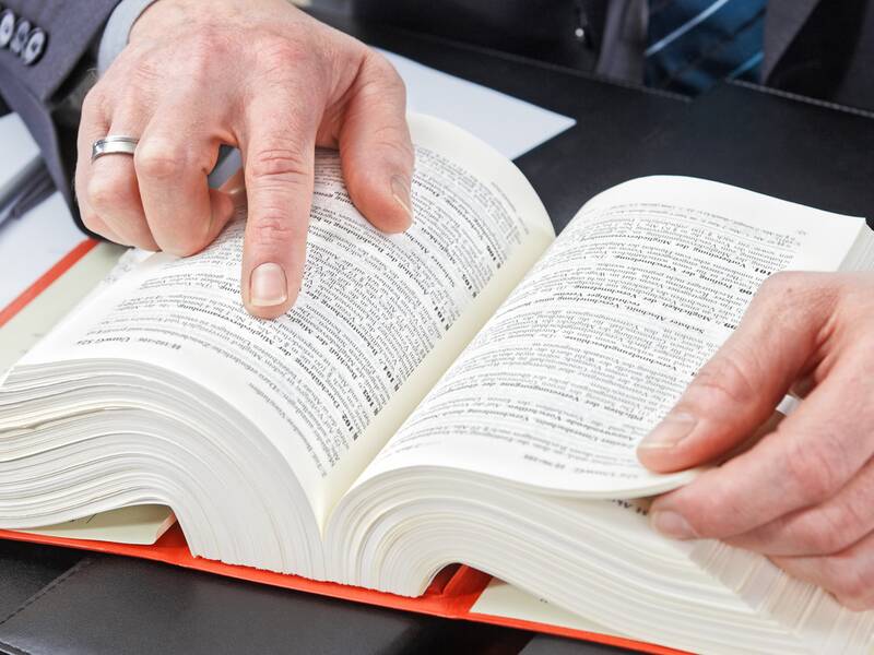 Ein deutsches Gesetzbuch liegt auf dem Tisch, ein Mann blättert darin und zeigt auf einen Paragrafen. 