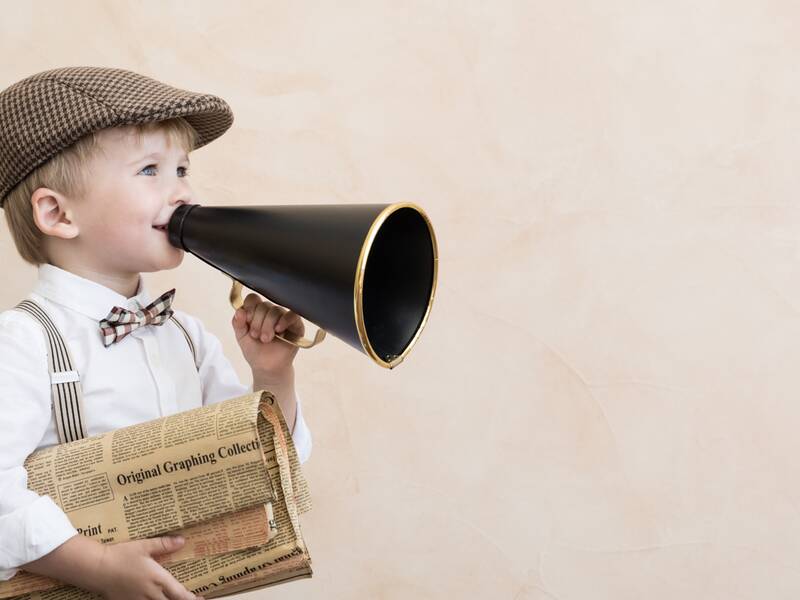 Symbolbild: Kind, dass eine Mütze, Fliege, ein weißes Hemd und Hosenträger trägt, hält ein altmodisches Megaphone in der einen und eine alte Zeitung in der anderen Hand. 