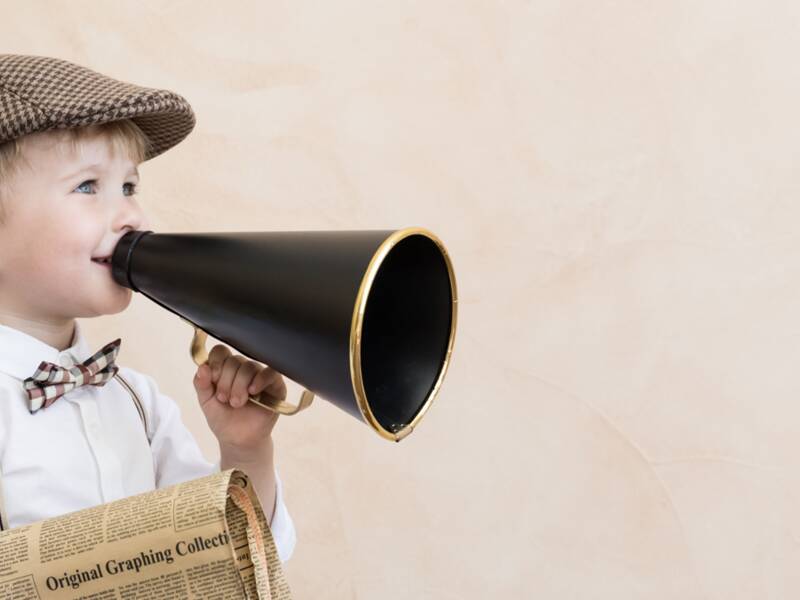 Symbolbild: Kind, dass eine Mütze, Fliege, ein weißes Hemd und Hosenträger trägt, hält ein altmodisches Megaphone in der einen und eine alte Zeitung in der anderen Hand. 