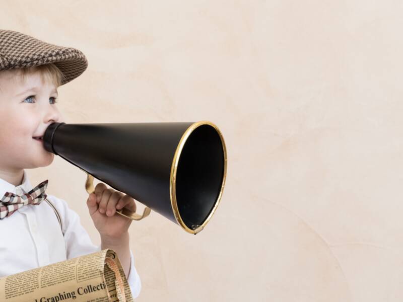 Symbolbild: Kind, dass eine Mütze, Fliege, ein weißes Hemd und Hosenträger trägt, hält ein altmodisches Megaphone in der einen und eine alte Zeitung in der anderen Hand. 