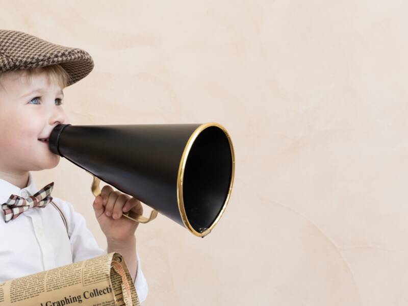 Symbolbild: Kind, dass eine Mütze, Fliege, ein weißes Hemd und Hosenträger trägt, hält ein altmodisches Megaphone in der einen und eine alte Zeitung in der anderen Hand. 