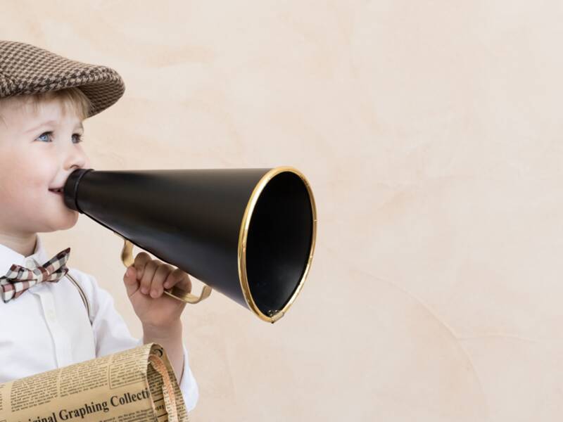 Symbolbild: Kind, dass eine Mütze, Fliege, ein weißes Hemd und Hosenträger trägt, hält ein altmodisches Megaphone in der einen und eine alte Zeitung in der anderen Hand. 