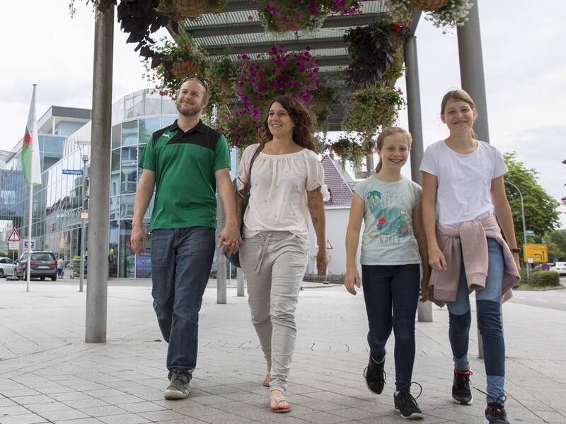 Eien Familie flaniert unter einer Galerie in der Fußgängerzone, im Hintergrund ein Einkaufszentrum und Fahnen