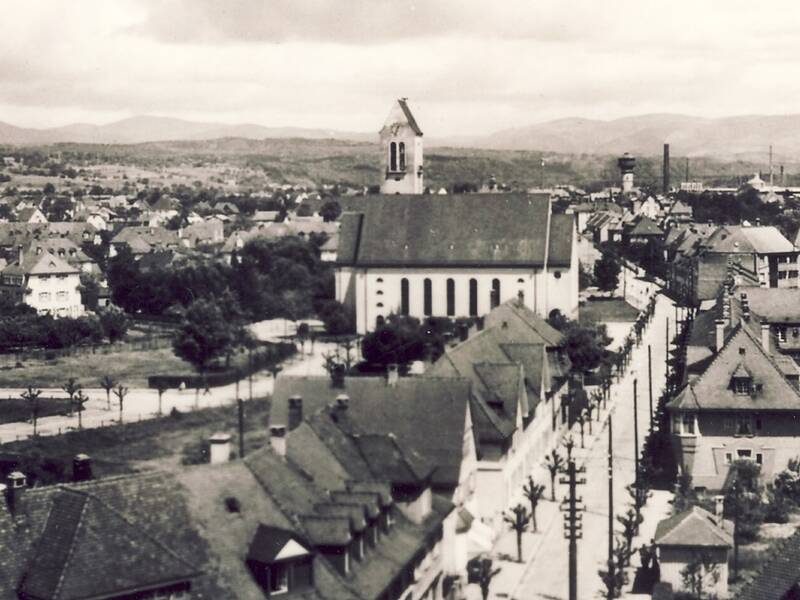 Die Schwarz-Weiß-Fotografie zeigt eine Stadtansicht Rheinfeldens aus den 1950er Jahren mit einer Kirche im Zentrum, umgeben von dicht bebaute Wohnhäusern und Straßen, im Hintergrund sind Hügel zu sehen.