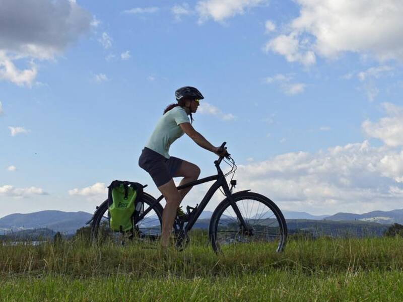Zwei Fahrradfahrer radeln einen Feldweg entlang, im Hintergrund Hügel und Berge