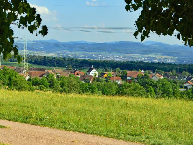 Blick von oberhalb auf das Dorf Nordschwaben. Im Vordergrund ist ein Teil eines Feldweges zu sehen, im Hintergrund sieht man das Tal des Hochrheins und Berge in der Schweiz. 