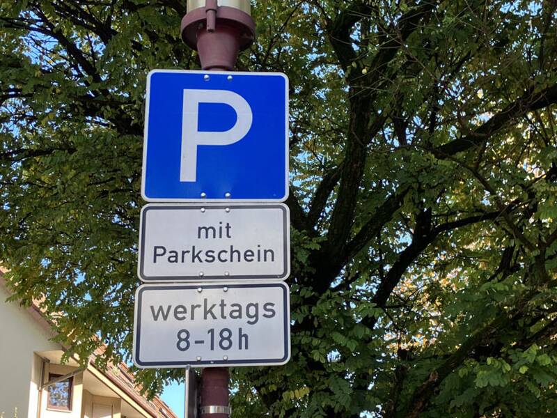 Verkehrsschild Parken mit Zusatzschildern "Parkschein" und "werktags von 8 - 18 Uhr". Im Hintergrund ist die noch grüne Krone eines Baumes zu sehen. 