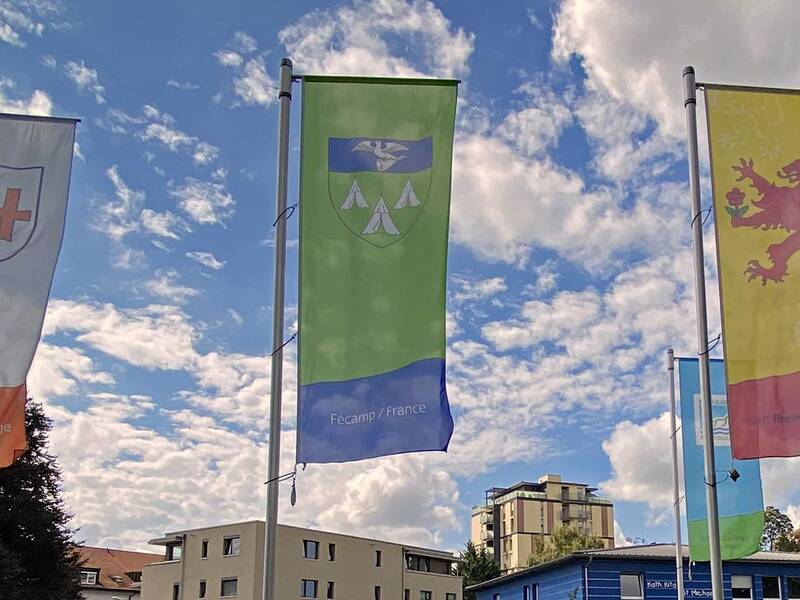 Fünf Fahnen, auf denen die Wappen der Rheinfelder Partnerstädte abgebildet sind, wehen im Wind. In Hintergrund blauer Himmel mit Wölkchen. 