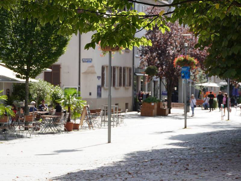 Eine belebte Fußgängerzone mit Cafés, Bäumen und Menschen, die auf der Straße und an Sitzbereichen verweilen.