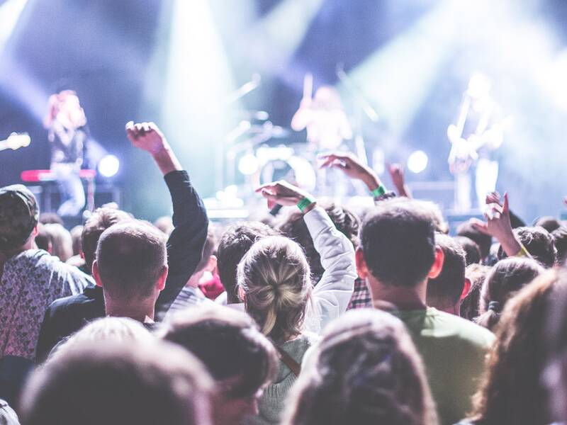 Publikumsperspektive bei einem Konzert: Die Künstler auf der Bühne sind nur angedeutet, im Vordergrund stehen die Menschen, die das Konzert verfolgen.   