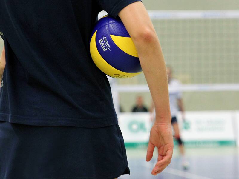 Hallensport: Im Vordergrund sieht man angedeutet eine Person, die einen blau-gelben Ball unter dem Arm hält. Im Hintergrund sind ein Volleyballnetz und weitere Spielerinnen zu erkennen. 