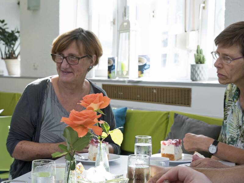 Eine Gruppe von Menschen sitzt an einem Tisch in einem hellen Raum und unterhält sich, während Getränke und ein Blumenstrauß in der Mitte des Tisches stehen.