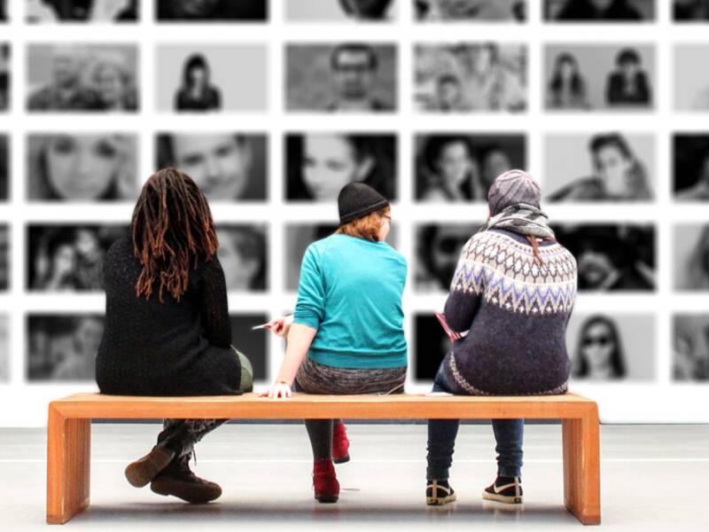 Drei Personen sitzen auf einer Bank in einer Ausstellung, die im Hintergrund zu sehen ist und verschiedene Porträts zeigt. 