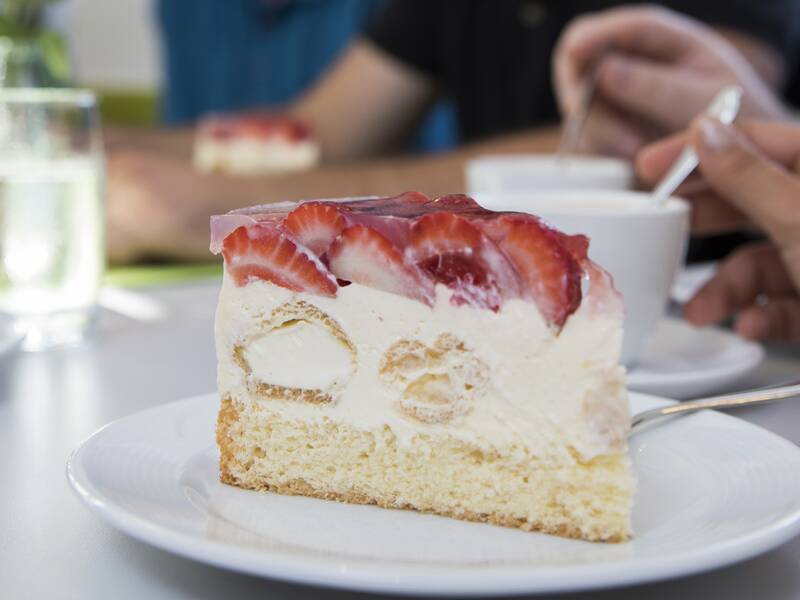 Auf dem Tisch sieht man im Vordergrund einen weißen Teller mit einem Stückchen Erdbeertorte stehen. Im Hintergund sieht man die Hände und Arme von Menschen sowie Kaffeetassen.