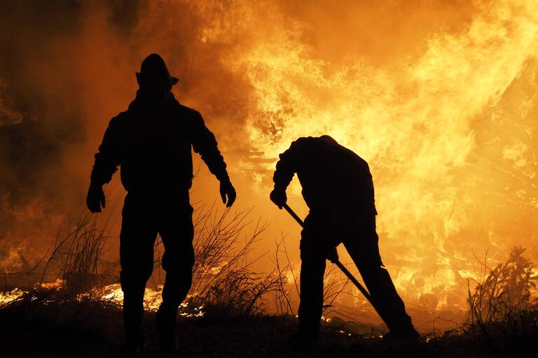 Zwei Männer, die nur als Schatten zu sehen sind, bekämpfen ein großes Feuer in der Natur. 