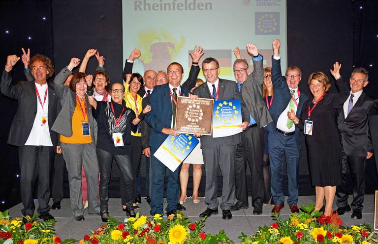 Eine Gruppe von Menschen auf einer Bühne, darunter Oberbürgermeister Klaus Eberhardt und Stadtammann Franco Mazzi, freut sich über die erhaltene Gold-Auszeichung bei dem Wettbewerb Entente Florale Europe. 