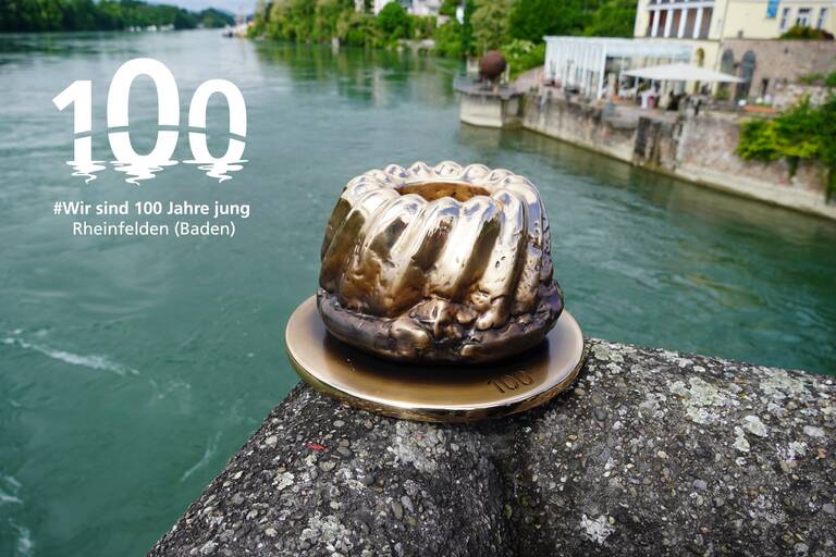 Kunstwerk "Kuchen und Zahl": Ein goldener Gugelhupf auf einem runden Tablett mit der Zahl 100, der auf einer Ecke des Geländers der alten Rheinbrücke steht. Oben links im Bild das Jubiläumslogo. 