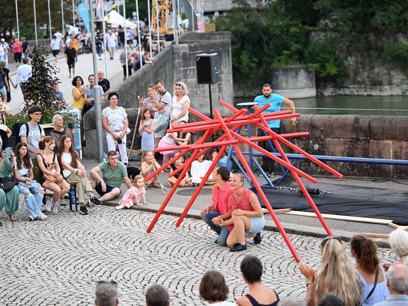 Ein Akrobatenpaar unter einer großen Mikadostäbe-Skulptur auf der alten Rheinbrücke