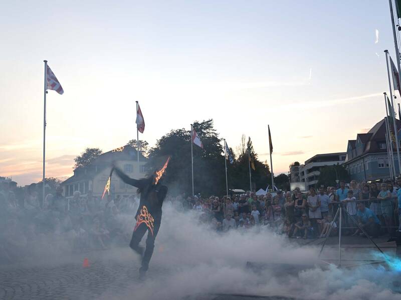 Brückensensationen 2026: Eine Person in einem Flammenanzug führt eine Feuershow vor einem Publikum auf, umgeben von Rauch und Flaggen im Hintergrund, während die Sonne untergeht.