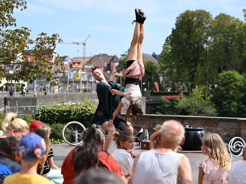 Brückensensationen 2026: Eine Artistin hebt eine andere Person in die Luft, während eine Gruppe von Zuschauern im Freien zuschaut.