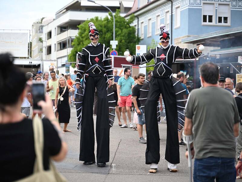 Brückensensationen 2026: Zwei aufwendig kostümierte Stelzenläufer unterhalten eine Menschenmenge auf einer belebten Straße.