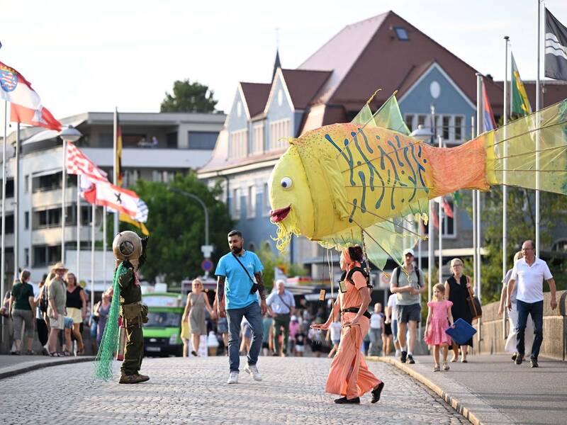 Brückensensationen 2026: Auf einer gepflasterten Brücke mit Flaggen und historischen Gebäuden im Hintergrund tragen Menschen in bunter Kleidung eine große, bunte Fischskulptur, während Passanten daneben gehen.
