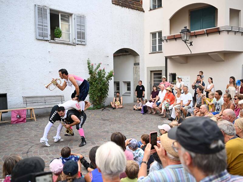 Brückensensationen 2026: Drei Akrobaten in bunter Kleidung führen in einem Innenhof vor einem sitzenden Publikum eine unterhaltsame, trompetenspielende Darbietung vor.