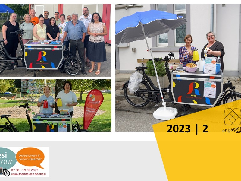 Eine Gruppe von Menschen steht an einem bunt gestalteten Fahrrad mit Informationsstand und Sonnenschirm, zusammen mit Bannern, die „Rhesi on Tour“ bewerben.