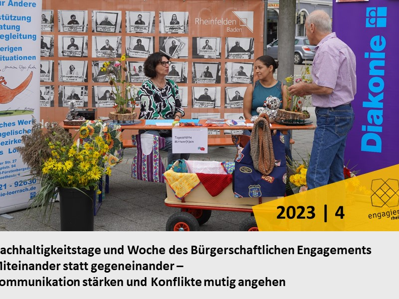 Ein Informationsstand mit zwei Personen, die sich unterhalten, ist von Blumen umgeben, und im Hintergrund befinden sich Banner der Diakonie sowie schwarz-weiße Porträts.