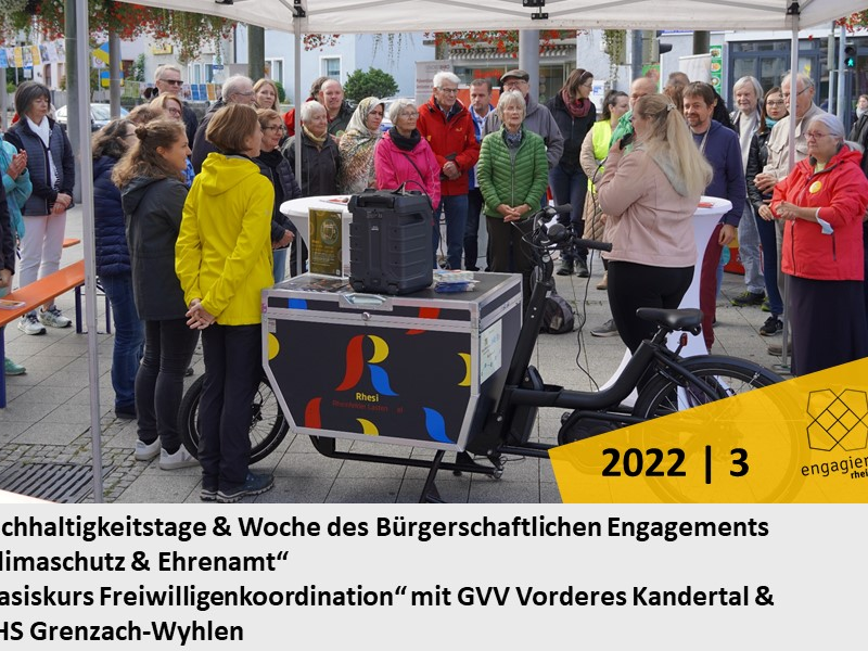 Eine Gruppe von Menschen hat sich unter einem Pavillon um ein Lastenfahrrad versammelt, während eine Person spricht; im Vordergrund sind Schilder mit Informationen zur Veranstaltung sichtbar.