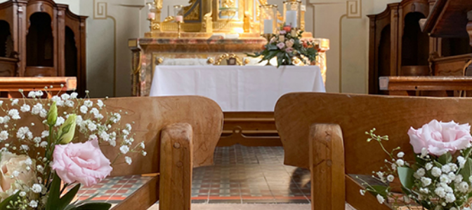 In einer Kirche sind die hölzernen Stuhlreihen mit Blumen geschmückt, die zu einem prunkvollen goldenen Altar führen, der mit weiteren floralen Arrangements dekoriert ist.