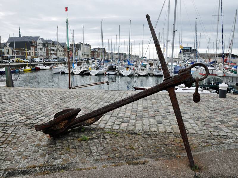 Hafen von Fécamp. Im Vordergrund liegt ein Anker auf Kopfsteinpflaster. Im Hintergrund sind Boote zu sehen. 