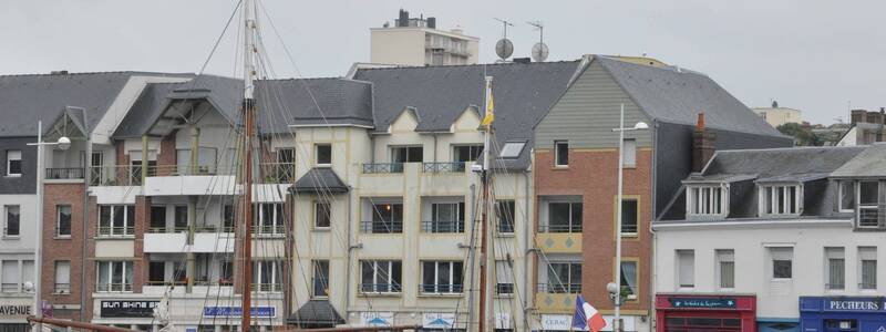 Ein blaues Segelschiff liegt am Ufer vor einer Reihe von mehrstöckigen Gebäuden, die überwiegend aus grauem und rotem Mauerwerk bestehen.