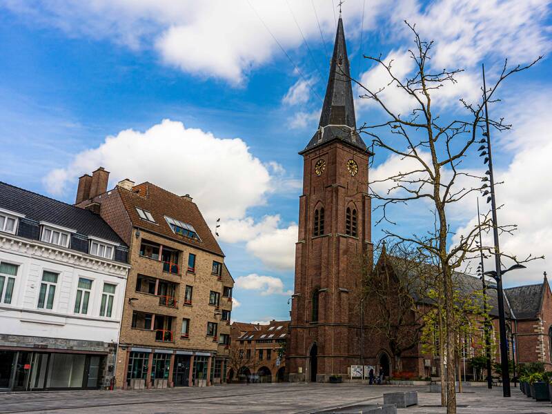 Auf dem Bild ist ein leerer Platz mit einem großen Kirchturm aus Backstein, umgeben von Wohnhäusern und einem blauen, wolkigen Himmel zu sehen.
