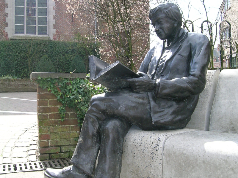Eine Bronzestatue eines sitzenden Mannes, der ein Buch liest, befindet sich auf einer Steinbank vor einer alten Kirche in einem gepflasterten Hof.