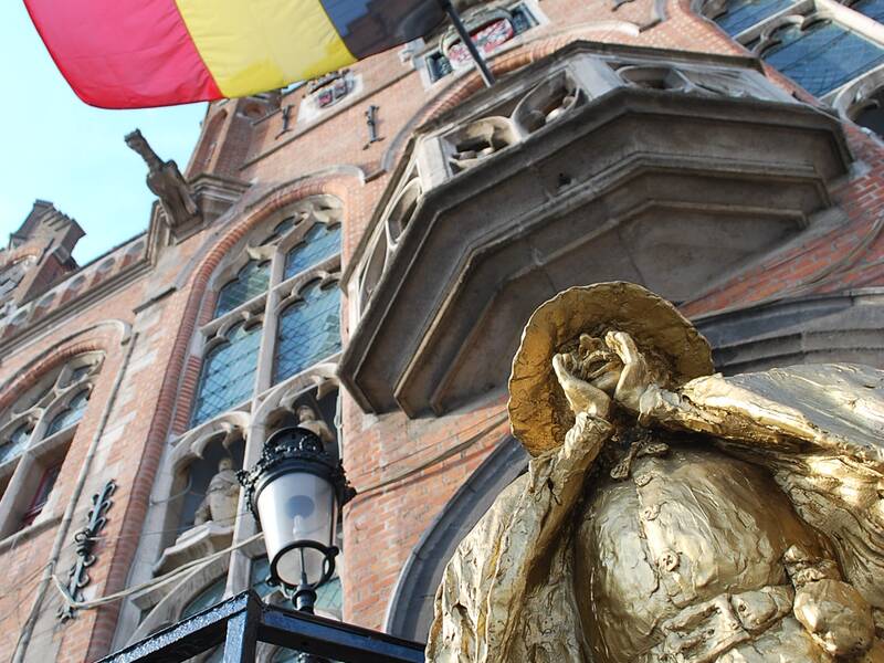Eine belgische Flagge weht über einer prächtigen, mit gotischen Elementen verzierten Backsteinfassade, neben einer goldenen Statue.