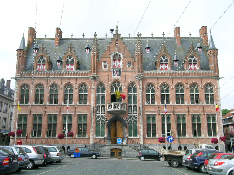 Ein großes, historisches Gebäude im gotischen Stil mit roten Ziegeln, zahlreichen verzierten Fenstern und einem zentralen Eingang, vor dem Autos geparkt sind.