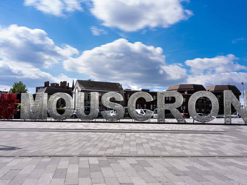 Ein großer, spiegelnder Schriftzug mit der Aufschrift "#MOUSCRON" steht auf einem gepflasterten Platz vor einem Himmel mit einigen Wolken.