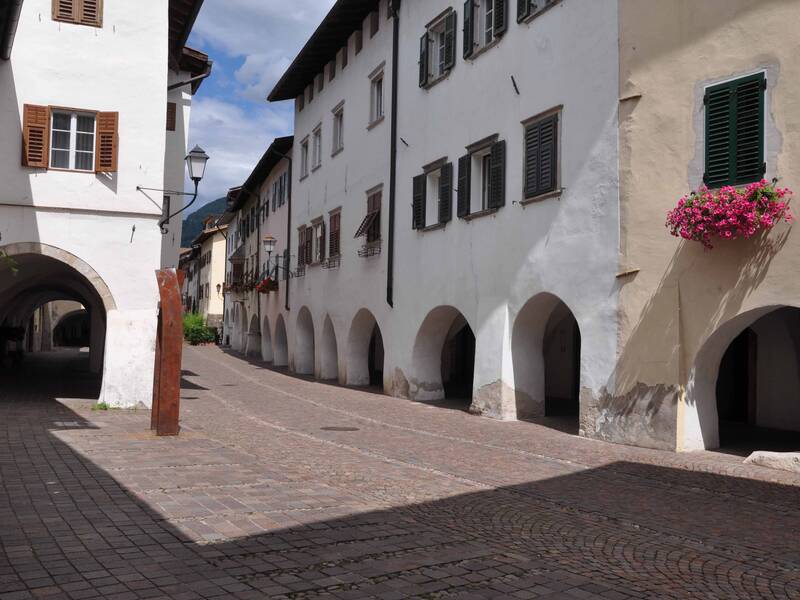 Eine gepflasterte Gasse mit Arkaden und historischen Gebäuden, an denen bunte Blumen an einem Fenster hängen.