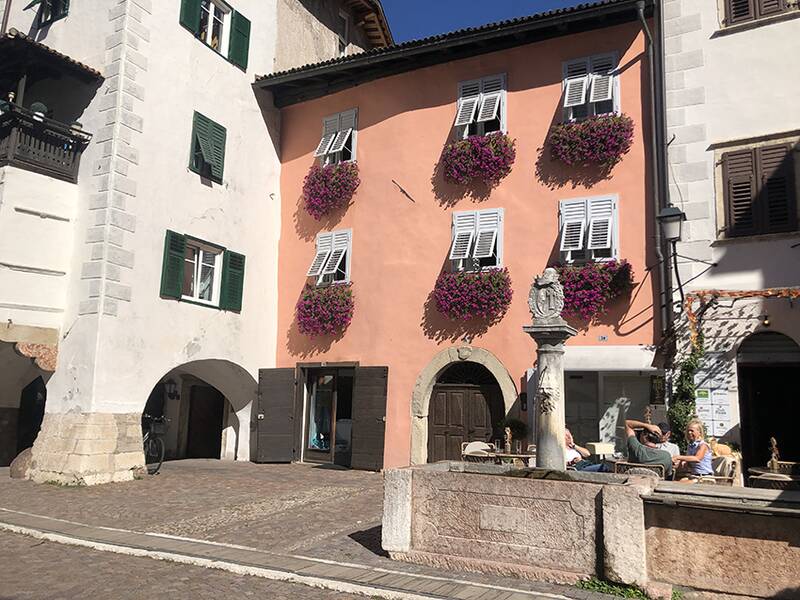 Ein rosafarbenes Gebäude mit weißen Fensterläden und üppigen Blumenkästen über den Fenstern, neben einem Brunnen, vor dem mehrere Personen sitzen.
