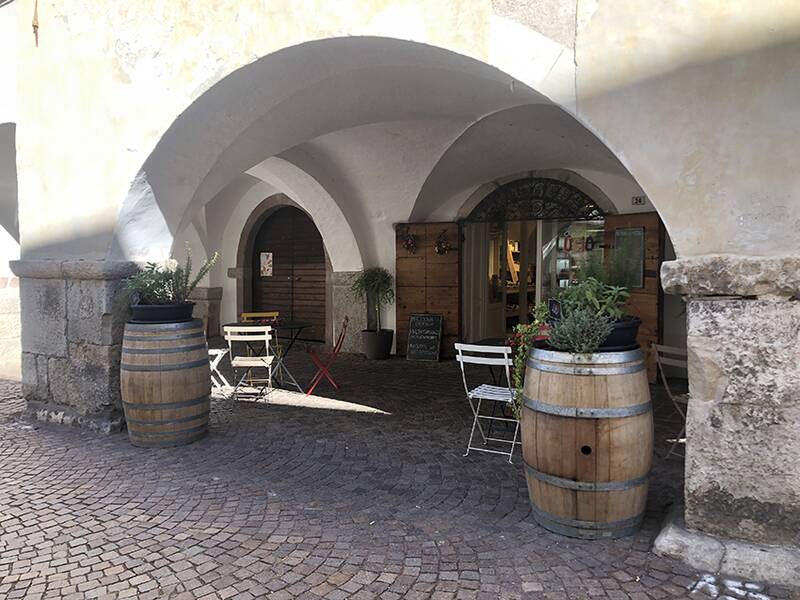 Unter einem steinernen Gewölbebogen steht ein kleines Café mit Tischen, Stühlen und Fässern, die als Pflanzgefäße dienen.