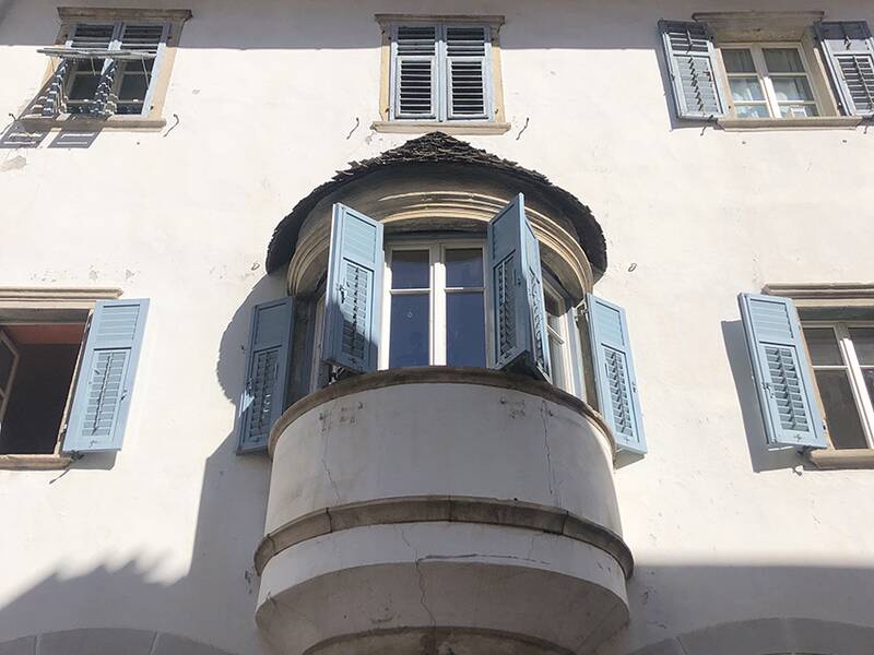Eine weiße Hausfassade mit blauen Fensterläden und einem hervorstehenden Erkerfenster in der Mitte.