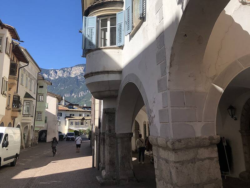 In einer sonnigen Altstadt mit historischen Gebäuden und Arkaden fährt ein Radfahrer die gepflasterte Straße entlang, während im Hintergrund Berge zu sehen sind.