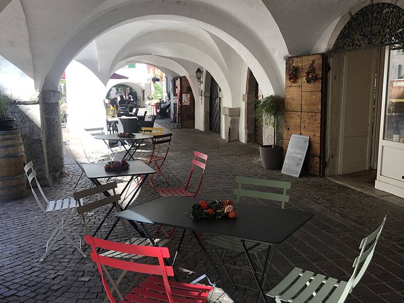 Unter einem steinernen Arkadengang stehen bunte Tische und Stühle, die zu einem Café gehören.