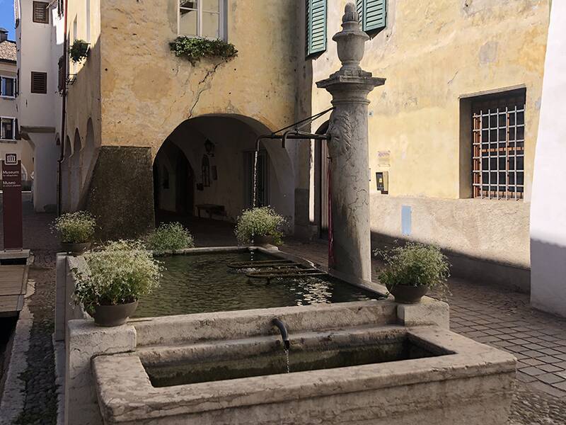 Ein steinerner Brunnen mit Pflanzenarrangements an den Ecken steht in einem Innenhof vor einem gelblichen Gebäude mit Arkaden und Fenstern mit grünen Fensterläden.