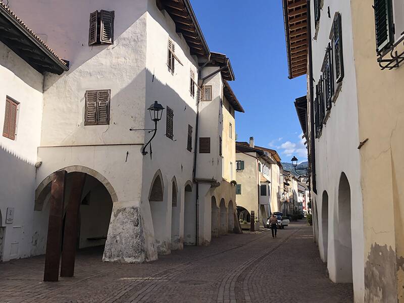 Eine schmale, gepflasterte Gasse mit historischen Gebäuden beiderseits, darunter Rundbögen und hölzerne Fensterläden, bei strahlend blauem Himmel.